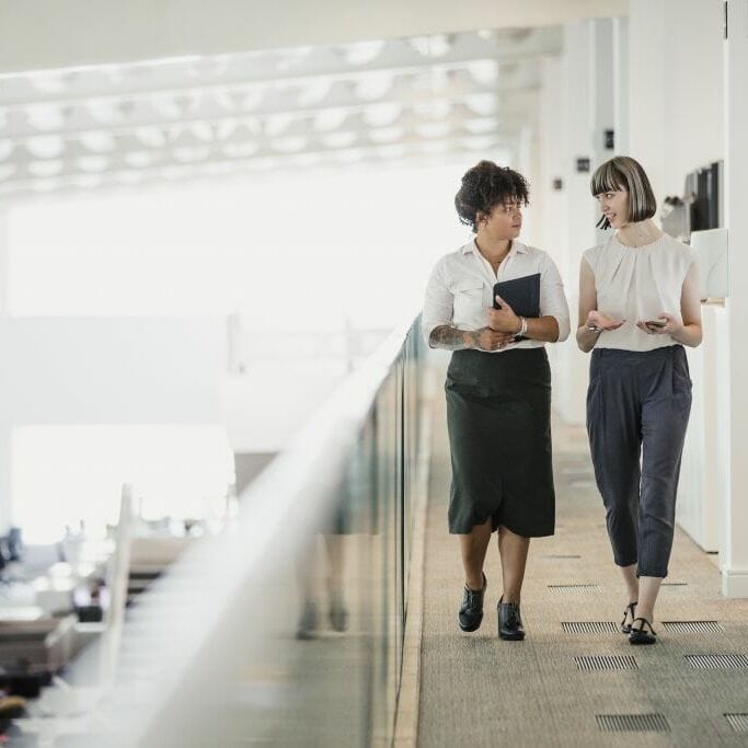 Two female professionals having a relaxed meeting while strolling along a raised walkway in an office auditorium.